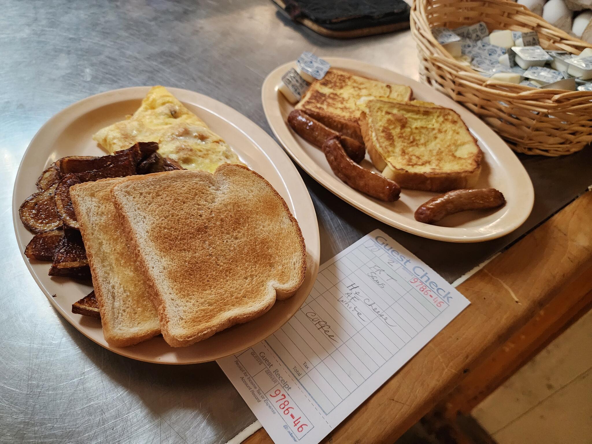 Two breakfast plates on the pass - a ham and cheese omelet with American fries and toast, and French toast with sausage links - beside a handwritten guest check