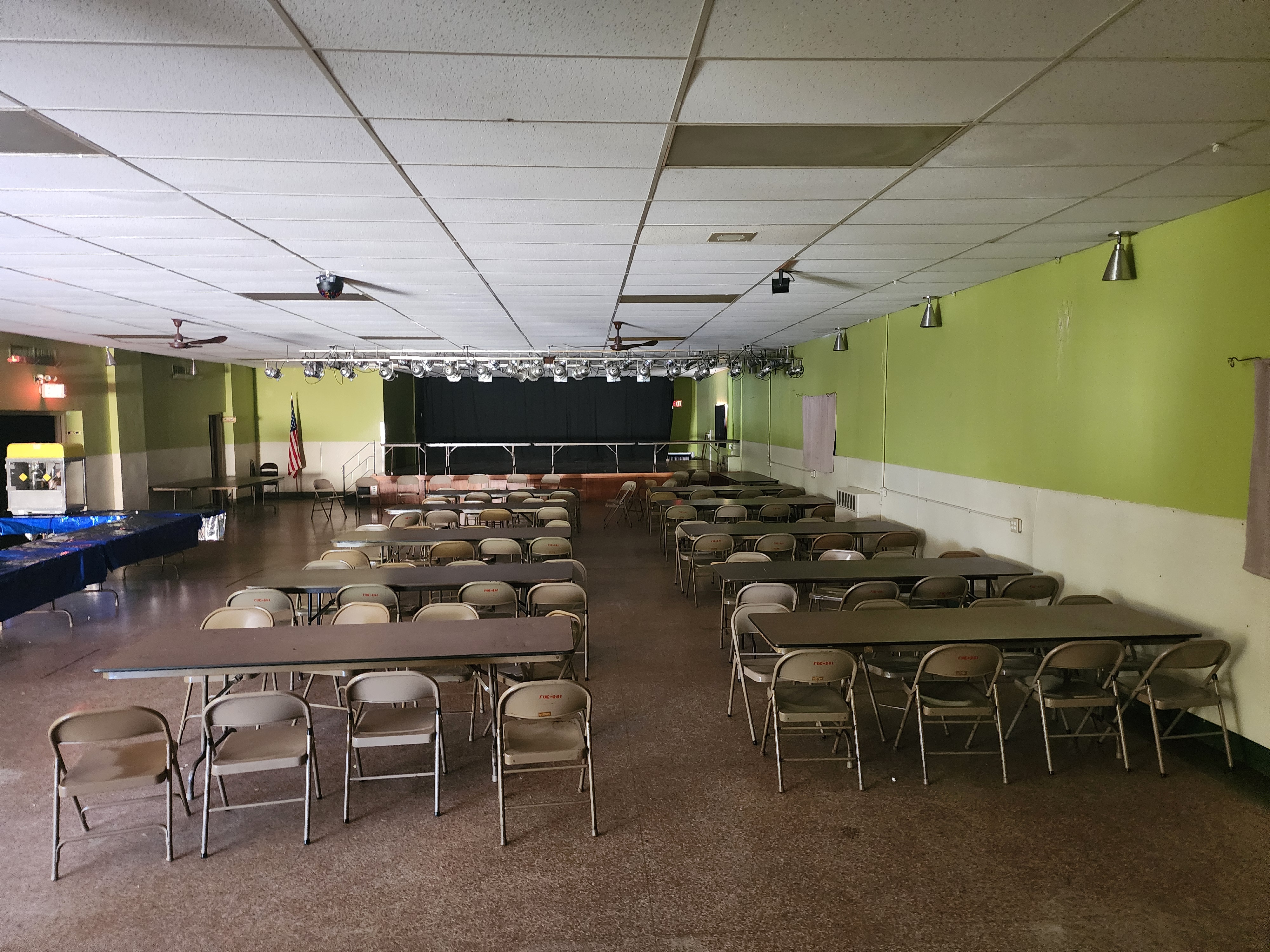 The upper hall from the back of the room, rows of tables and chairs facing the stage with the flag beside it